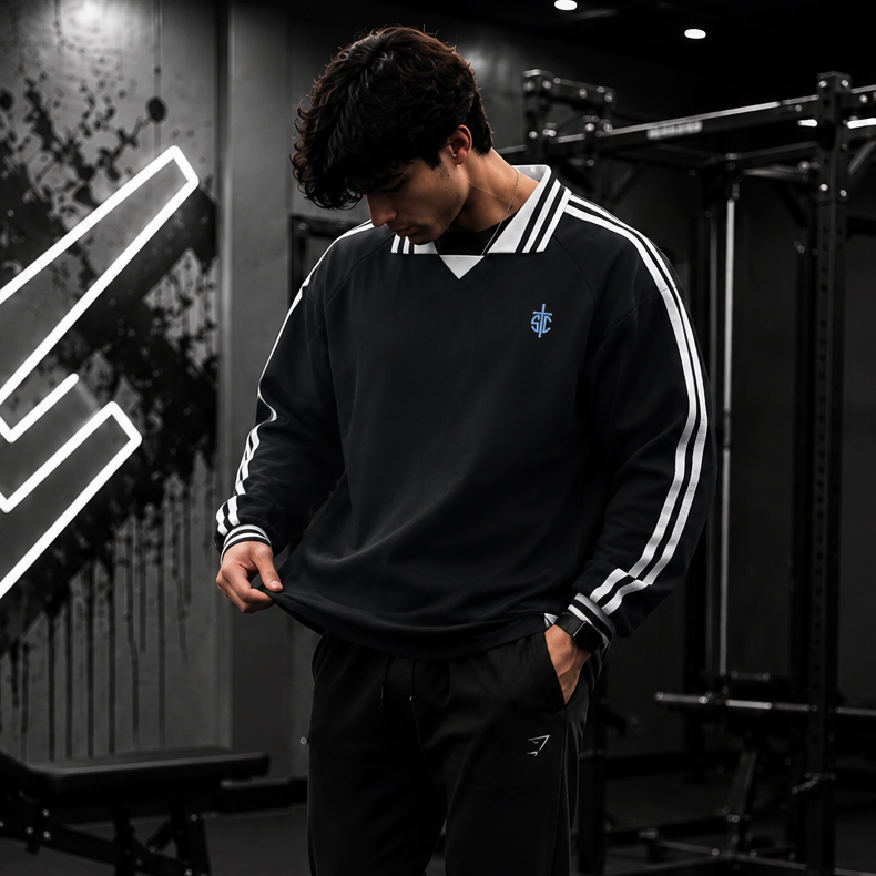 A man in a gym wearing a black long-sleeve sweatshirt with white stripes on the sleeves, black jogger pants, and white sneakers.