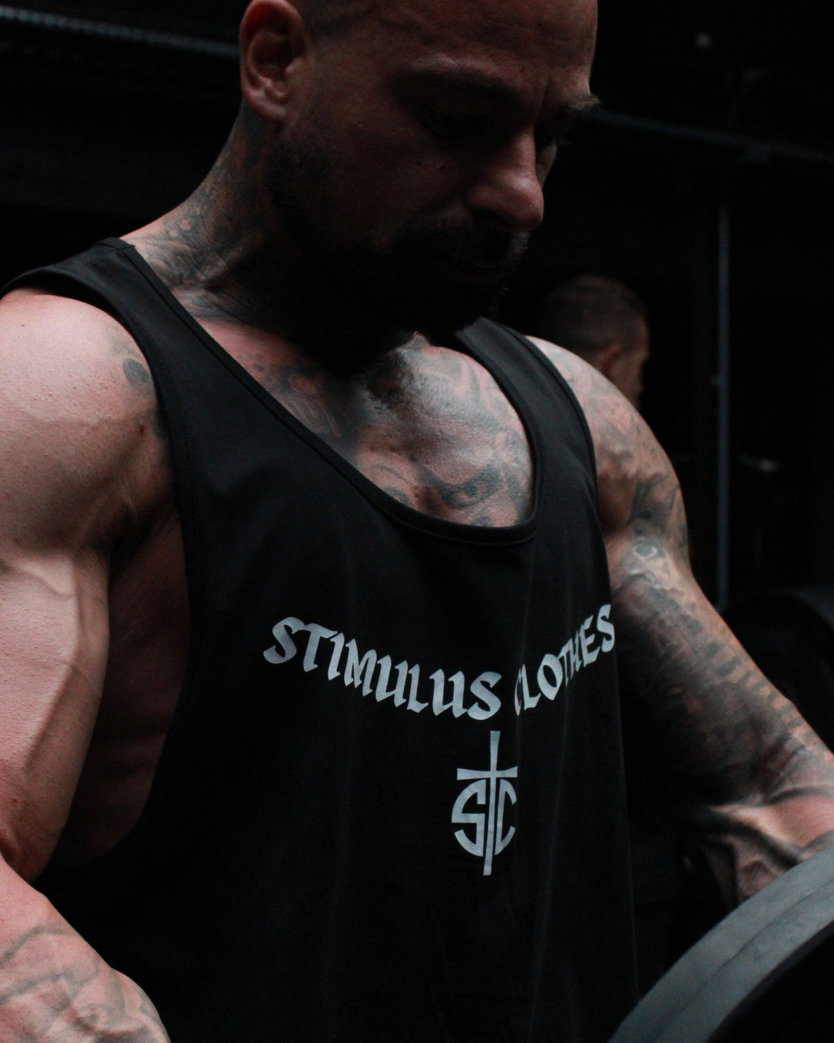 A muscular man with tattoos lifting a barbell in a dark gym setting, wearing a black tank top with the white text 'STIMULUS LONES' and a wrist wrap.