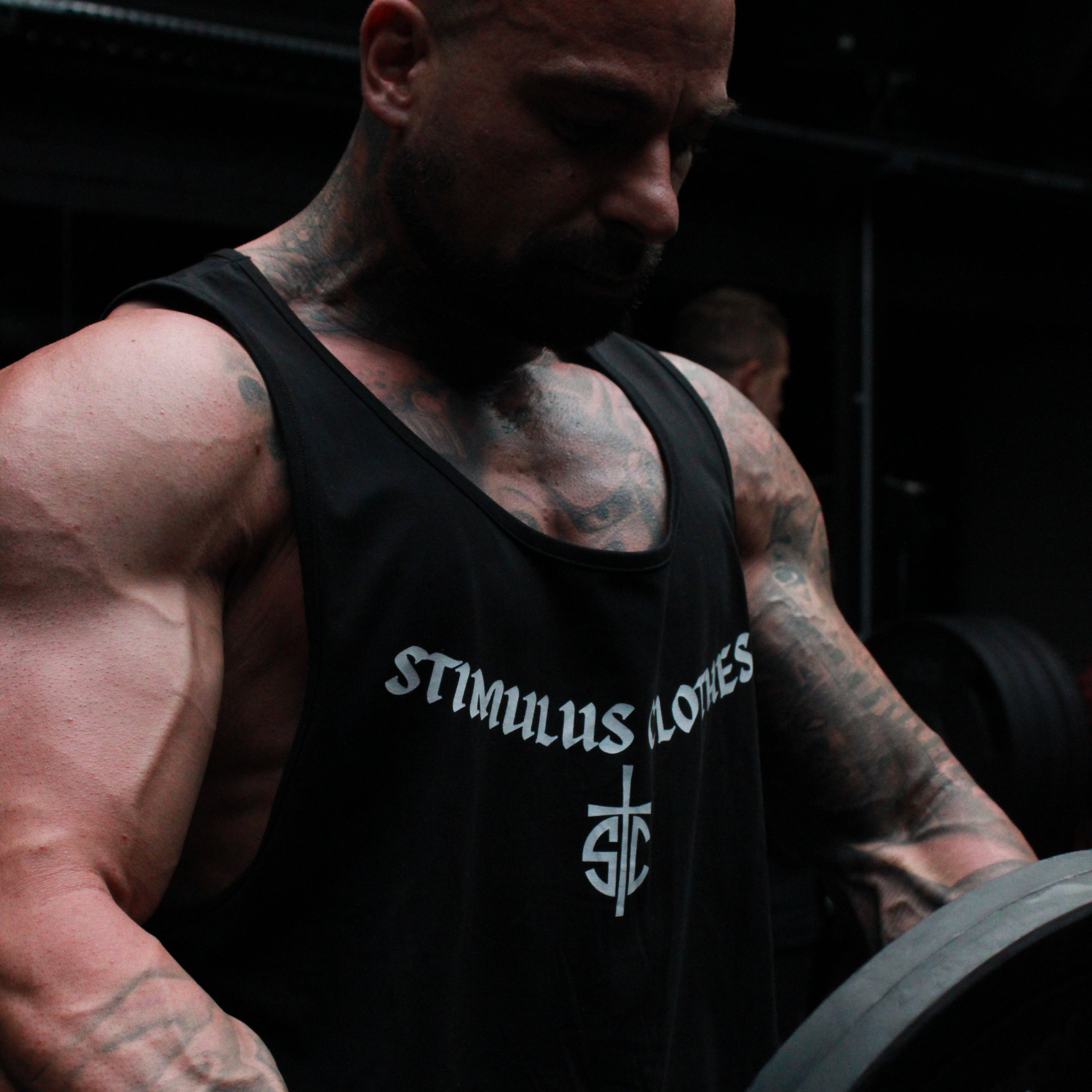 A muscular man with tattoos lifting a barbell in a dark gym setting, wearing a black tank top with the white text 'STIMULUS LONES' and a wrist wrap.