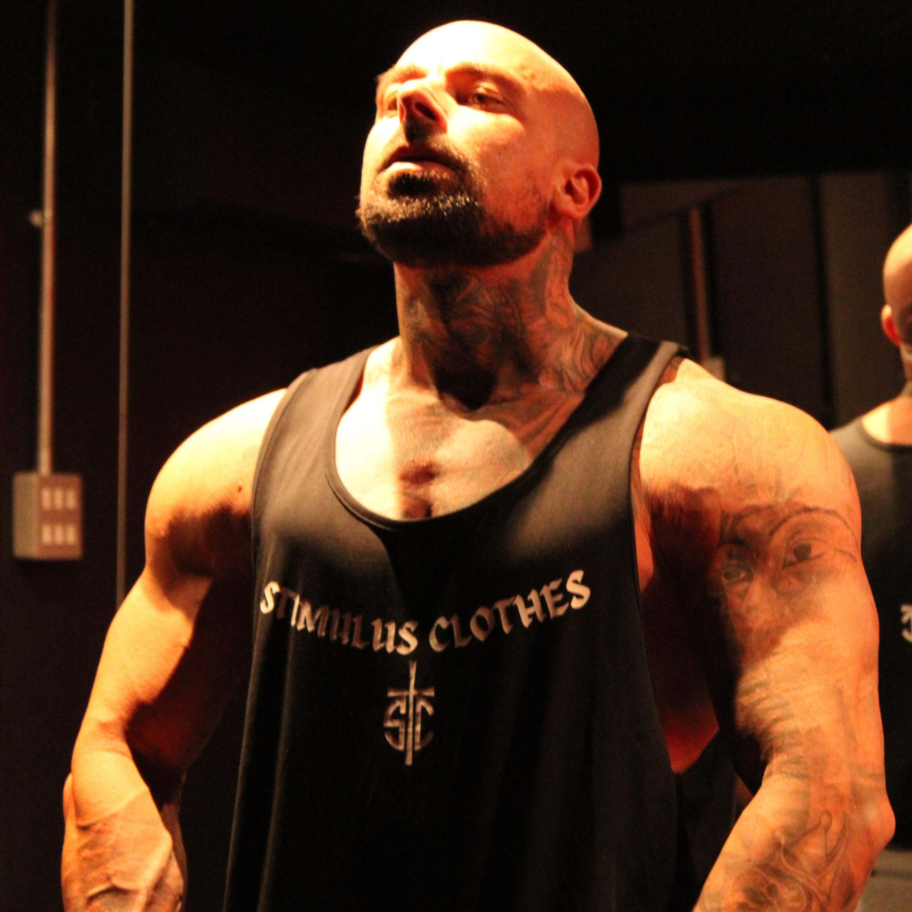 A muscular man wearing a black tank top with 'ST. INRIAH'S CLOTHES' printed on the chest, standing in a dimly lit indoor setting.