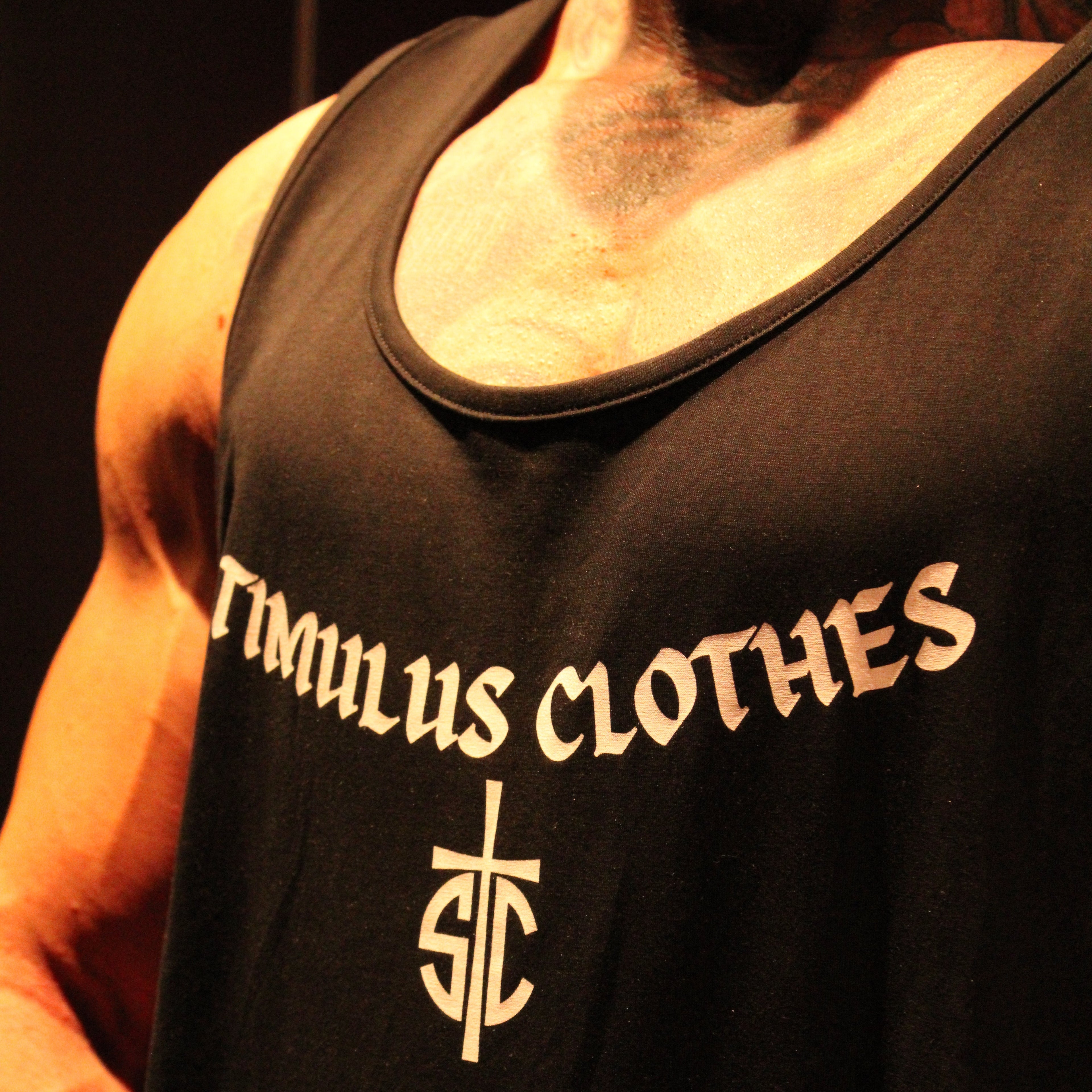 A close-up of a muscular person wearing a black tank top with the words 'TIMULUS CLOTHES' and a stylized 'TC' logo printed in a light beige or cream color on the chest.