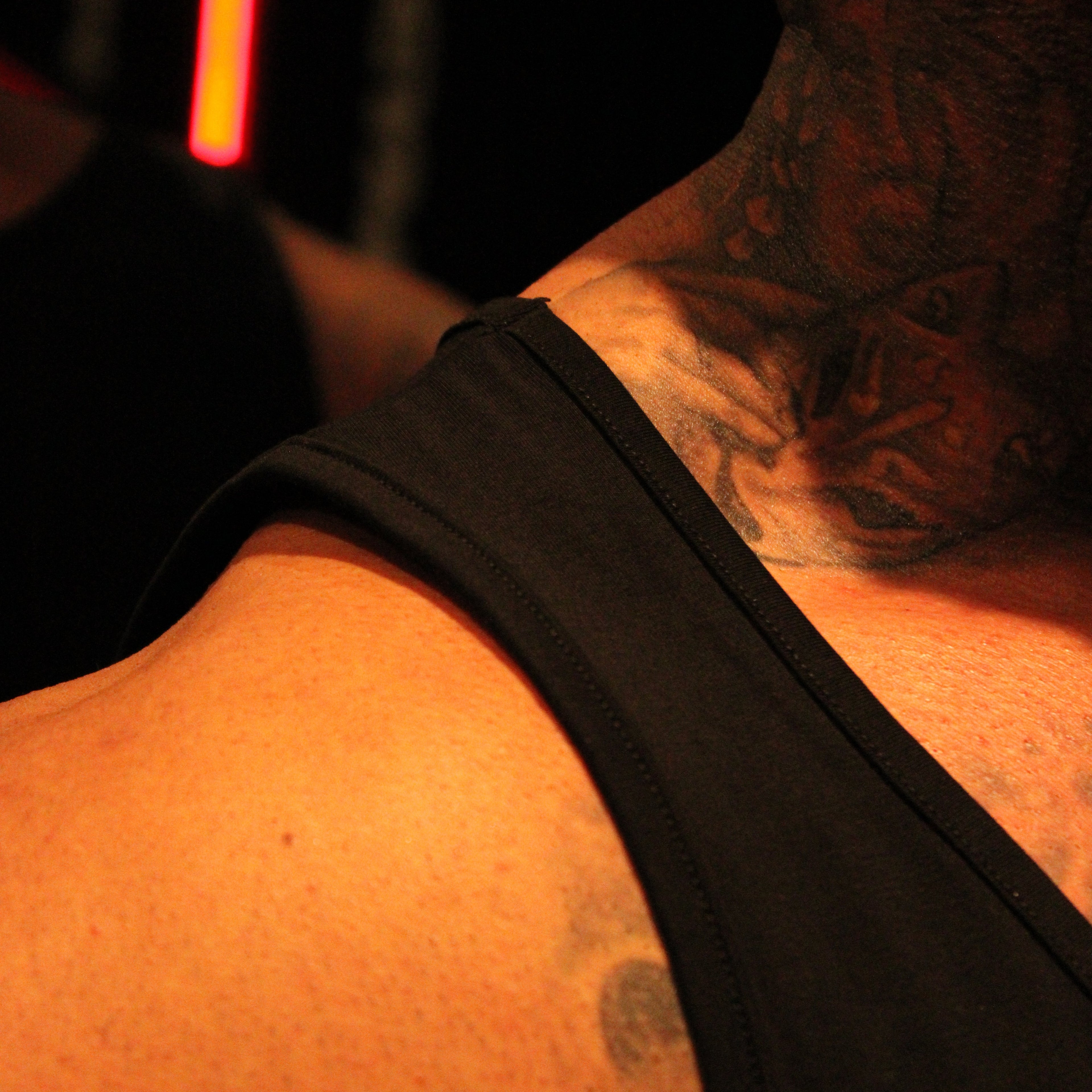 Close-up of a person's tattooed neck and shoulder wearing a black tank top, with warm orange lighting against a dark background.