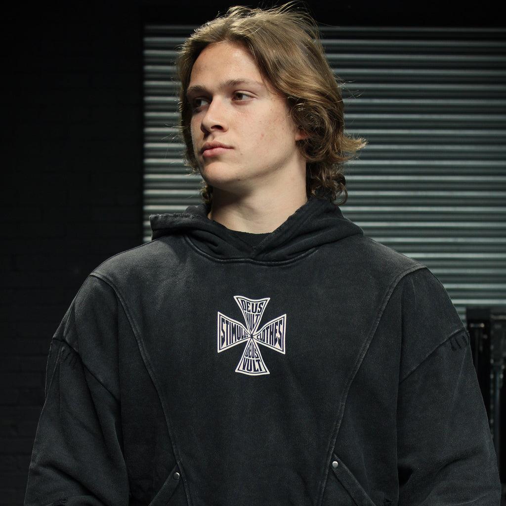 A man wearing a faded black pullover hoodie with a white and black Iron Cross graphic on the chest, standing against a dark background with horizontal window blinds.