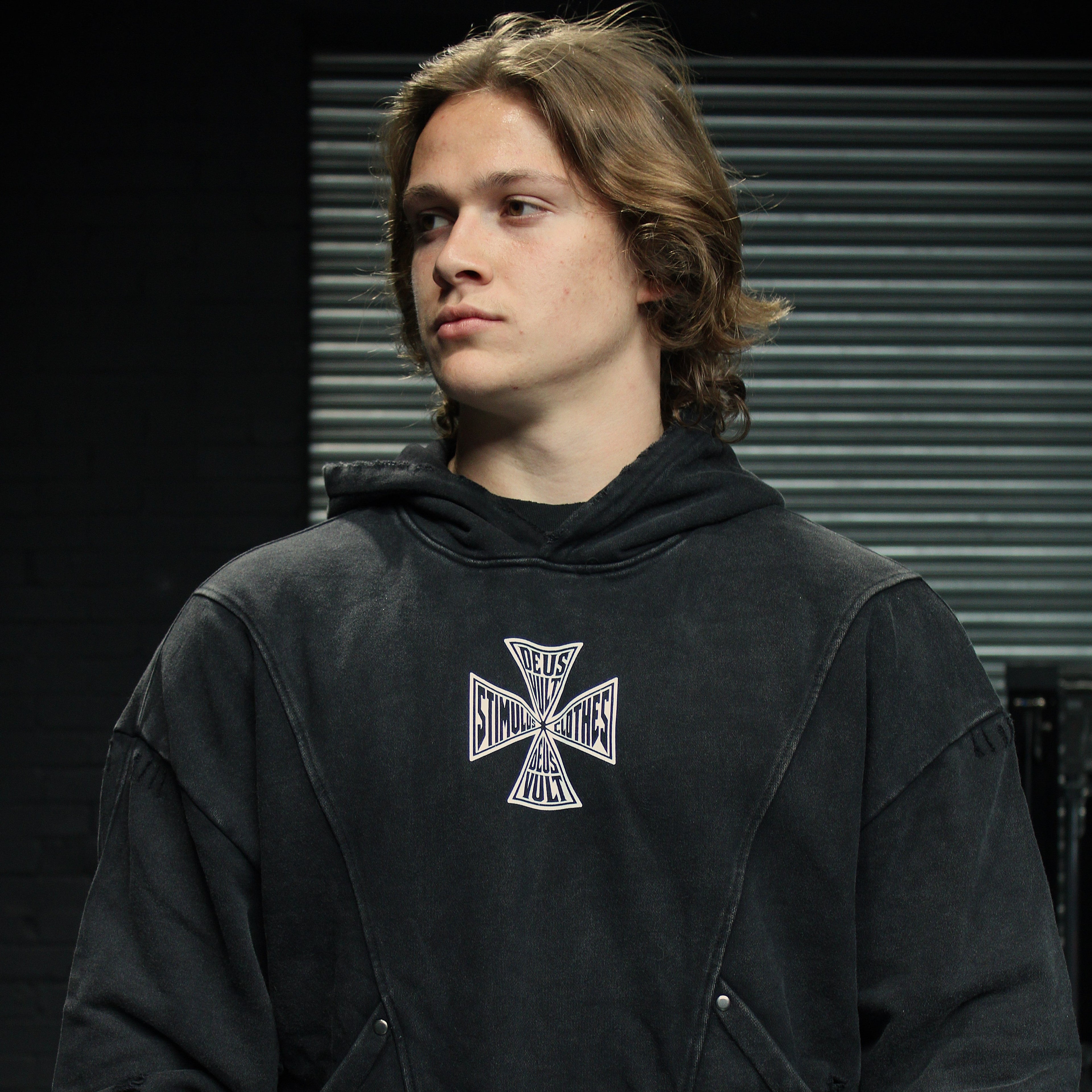 A man wearing a faded black pullover hoodie with a white and black Iron Cross graphic on the chest, standing against a dark background with horizontal window blinds.