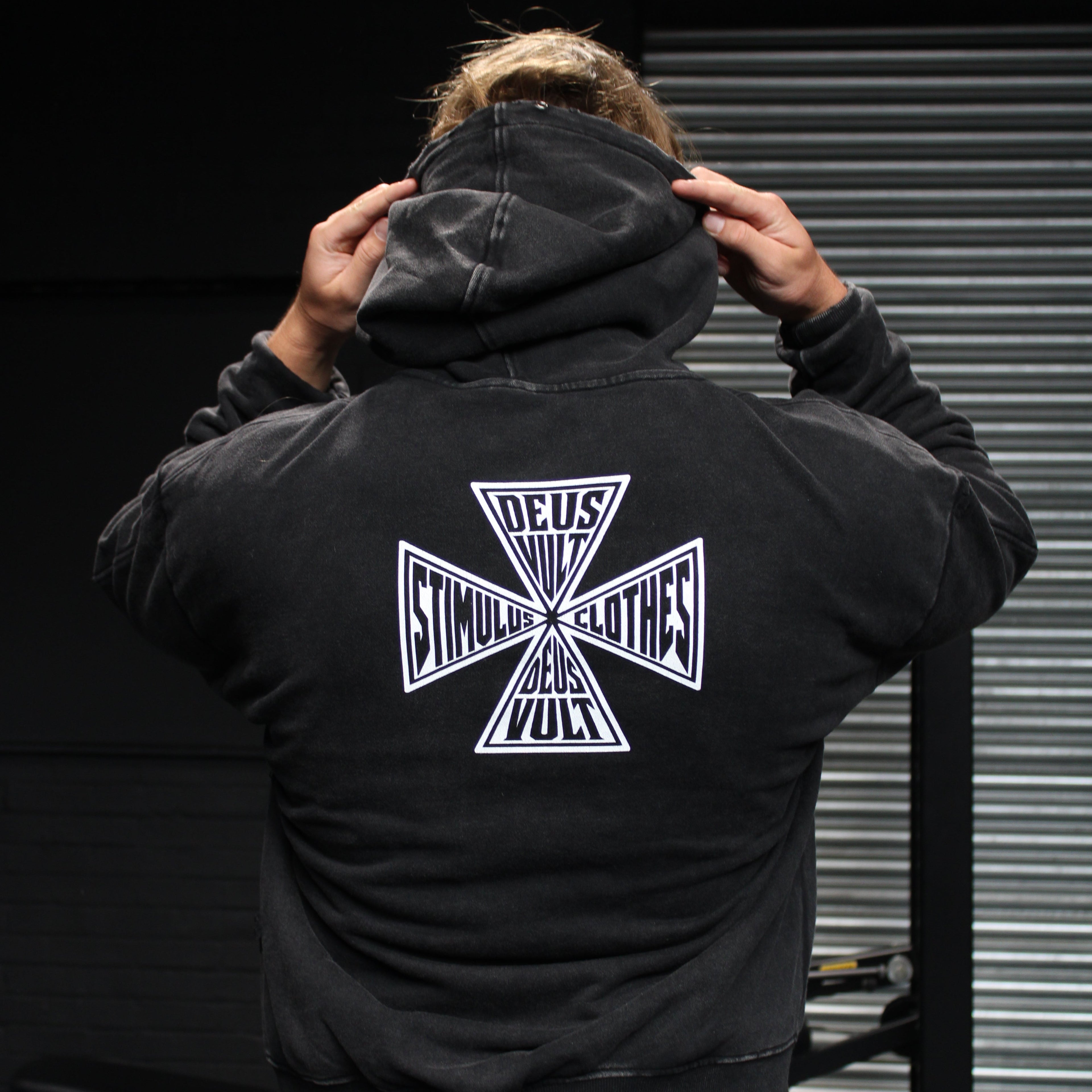 A person viewed from behind wearing a black hoodie with a large white graphic on the back, standing in a dark indoor setting with a metal roll-up door in the background.