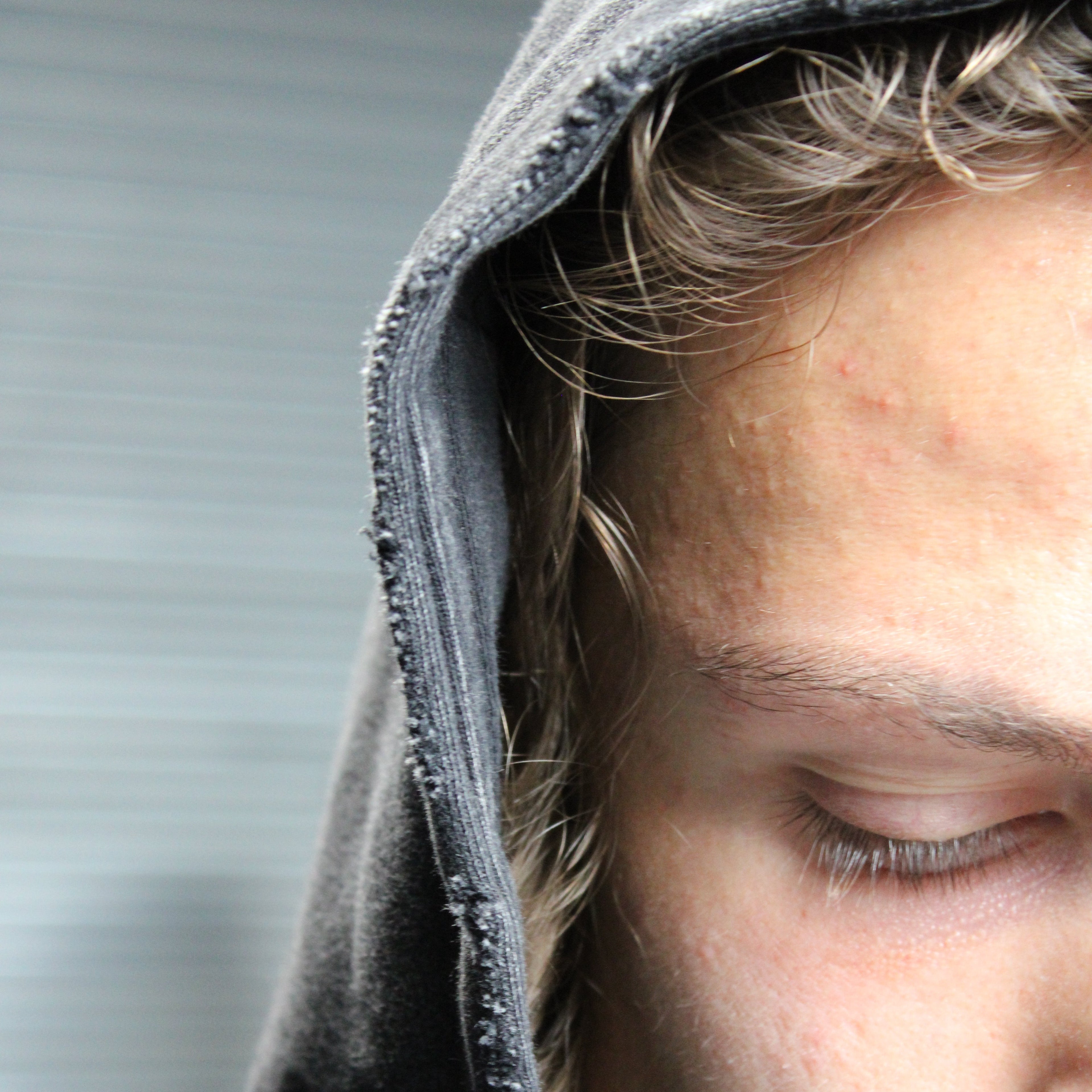 Close-up of a person wearing a dark gray hooded sweatshirt, with the hood pulled up and the person's face partially visible against a blurred gray background.