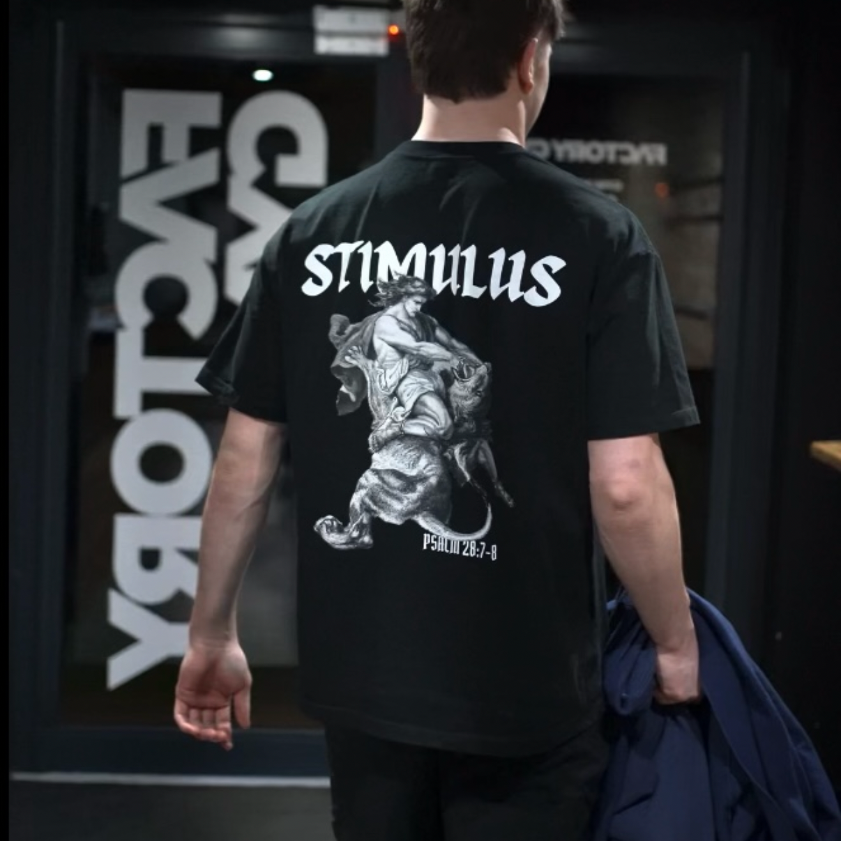 Person wearing a black t-shirt with 'STIMULUS' design and text, standing in a dark indoor setting.