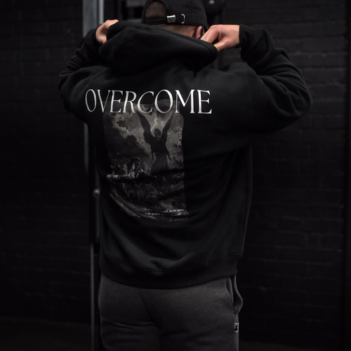 Person wearing a black hoodie with 'OVERCOME' printed on the back, standing against a dark brick wall.
