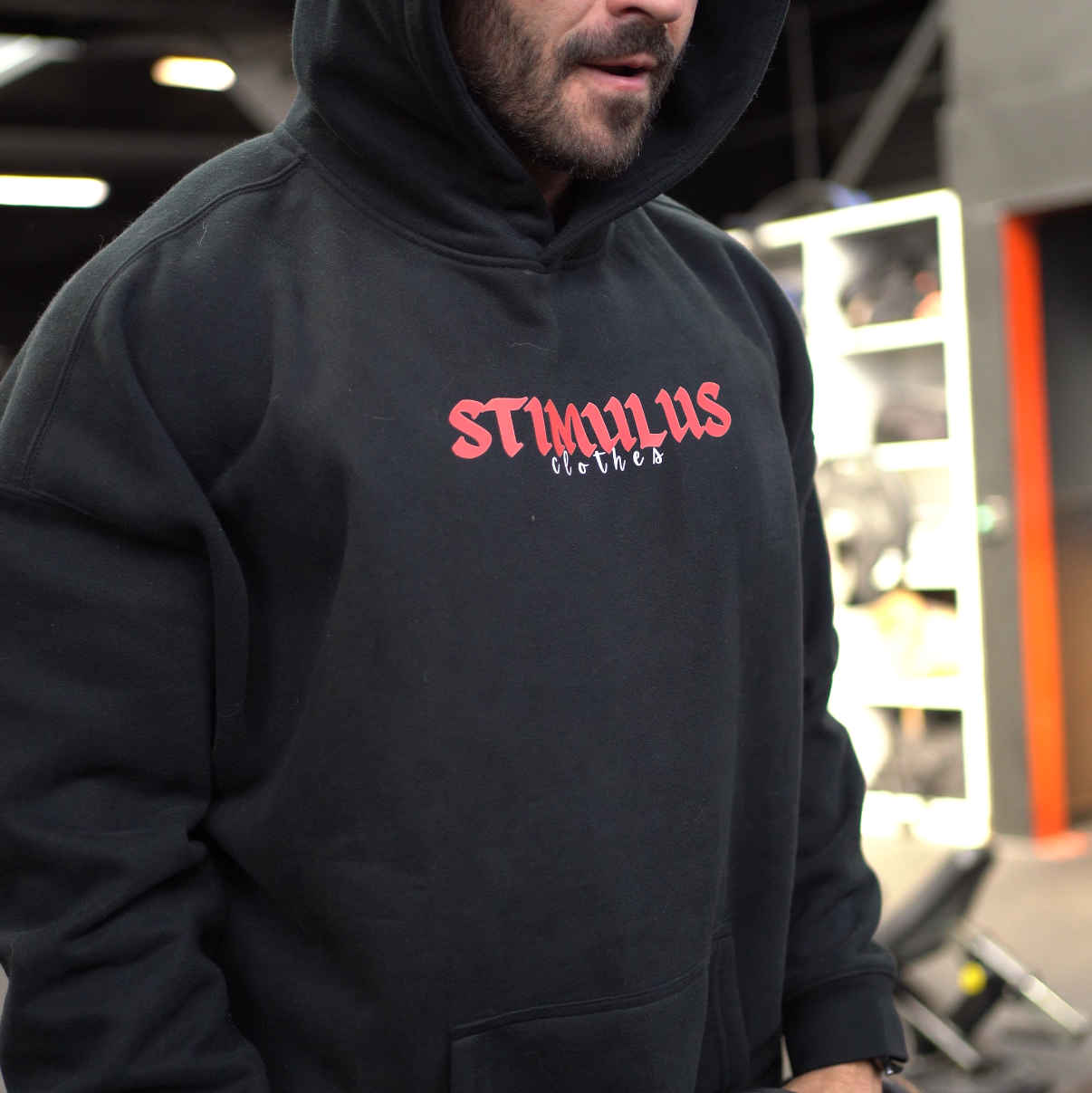 Person wearing a black hoodie with 'STIMULUS' branding, holding dumbbells in a gym setting.