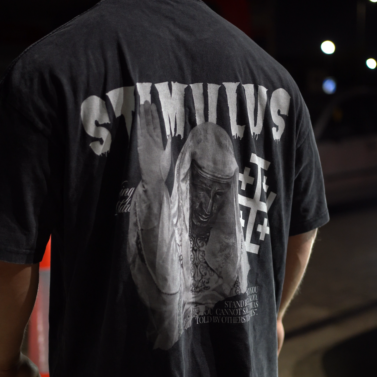 Person wearing a black t-shirt with a graphic design and text on the back, standing outdoors at night.