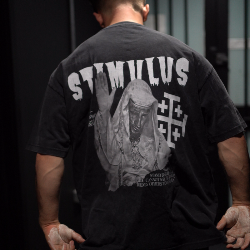 Person wearing a black t-shirt with 'STIMULUS' design on the back, standing in a dark indoor setting.