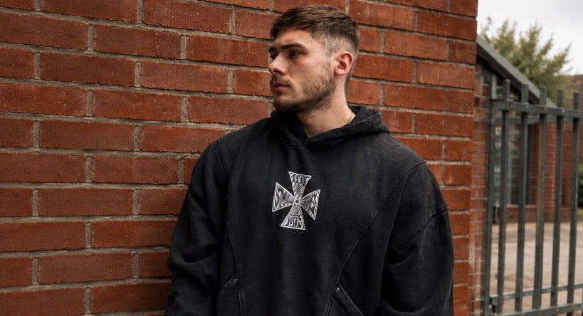 A man wearing a black pullover hoodie with a white Iron Cross graphic on the chest, standing against a red brick wall.