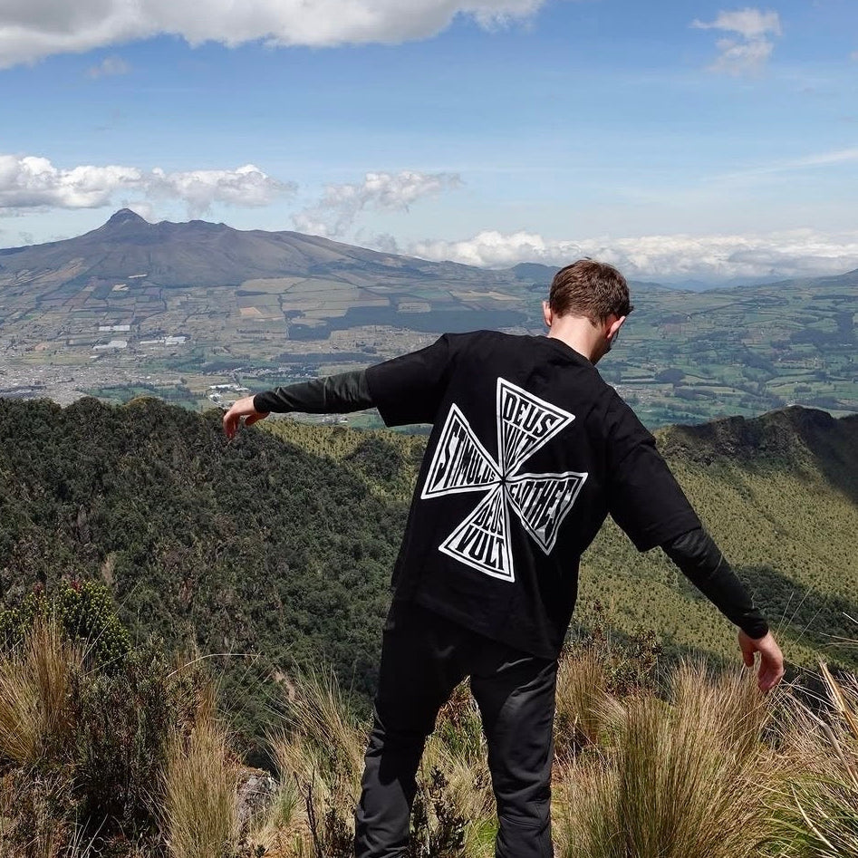 A man stands on a grassy mountain ridge with his back to the camera, wearing a black long-sleeve shirt with a large white graphic on the back that reads 'THIS WAY ONLY', black pants, and red shoes, overlooking a vast landscape under a cloudy blue sky.