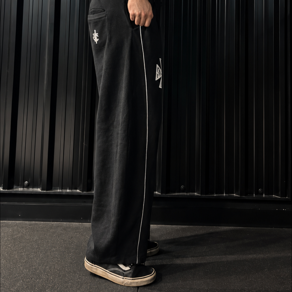 A man wearing black athletic track pants with a white side stripe and a black tank top, standing in a gym in front of a black corrugated metal wall.