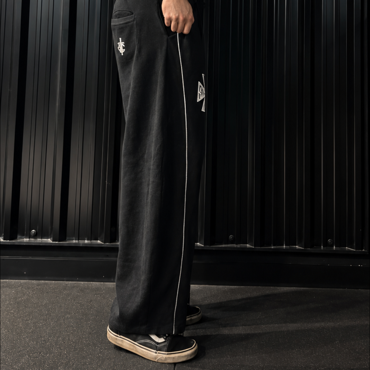 A man wearing black athletic track pants with a white side stripe and a black tank top, standing in a gym in front of a black corrugated metal wall.