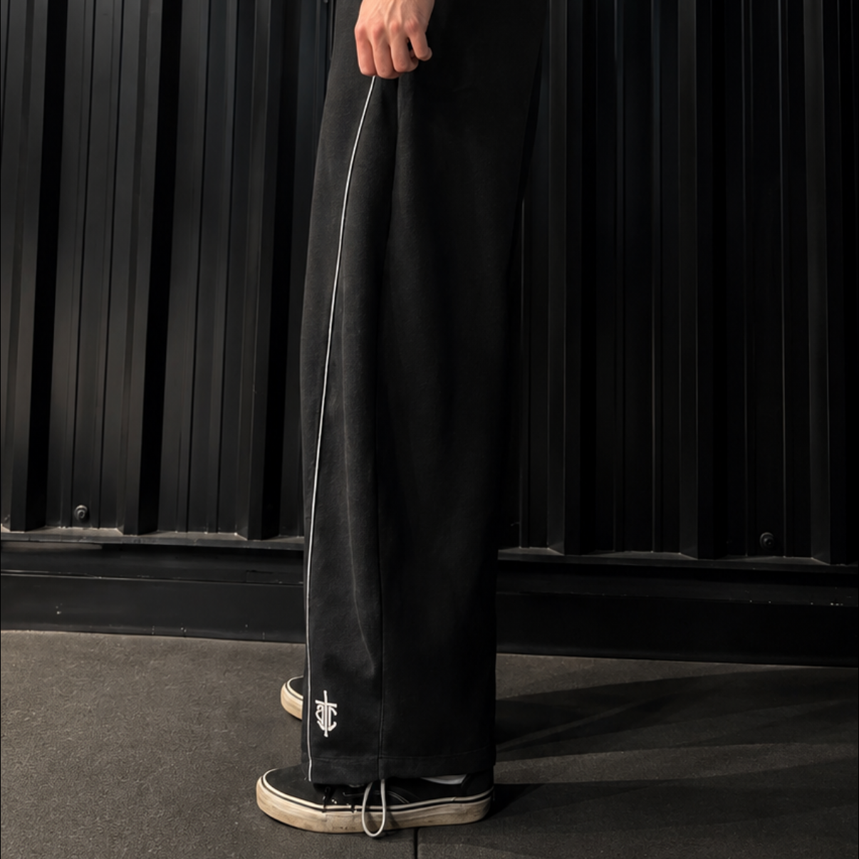 A man wearing a black sleeveless tank top and matching black wide-leg trousers stands in profile against a dark corrugated metal wall, looking down. The product appears to be the black pants, with a thin light-colored stripe running down the side.