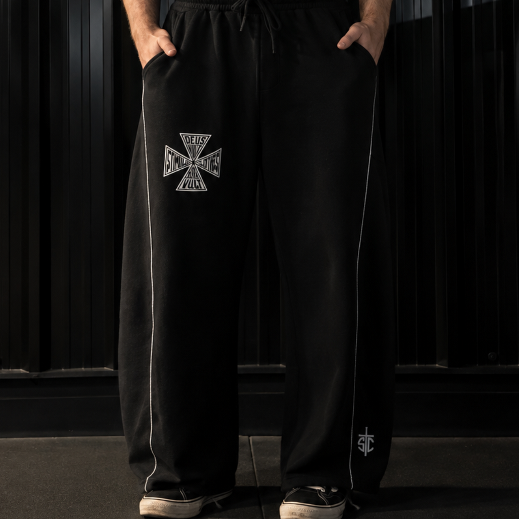 A man stands in a dark studio wearing a black sleeveless tank top with the word 'stimulus' in white across the chest and black drawstring sweatpants with a white Iron Cross emblem on the left leg.