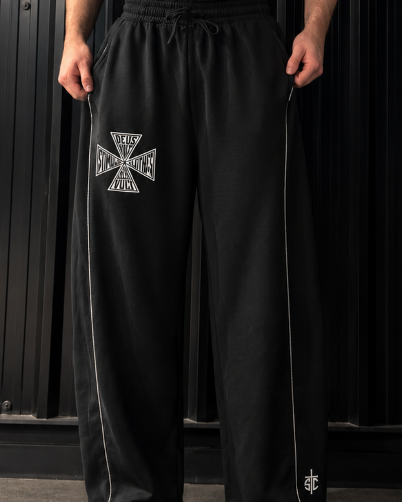 A man wearing a black tank top with 'stimulus' printed on the chest and black sweatpants with a silver Iron Cross emblem on the thigh, standing against a black corrugated metal backdrop.