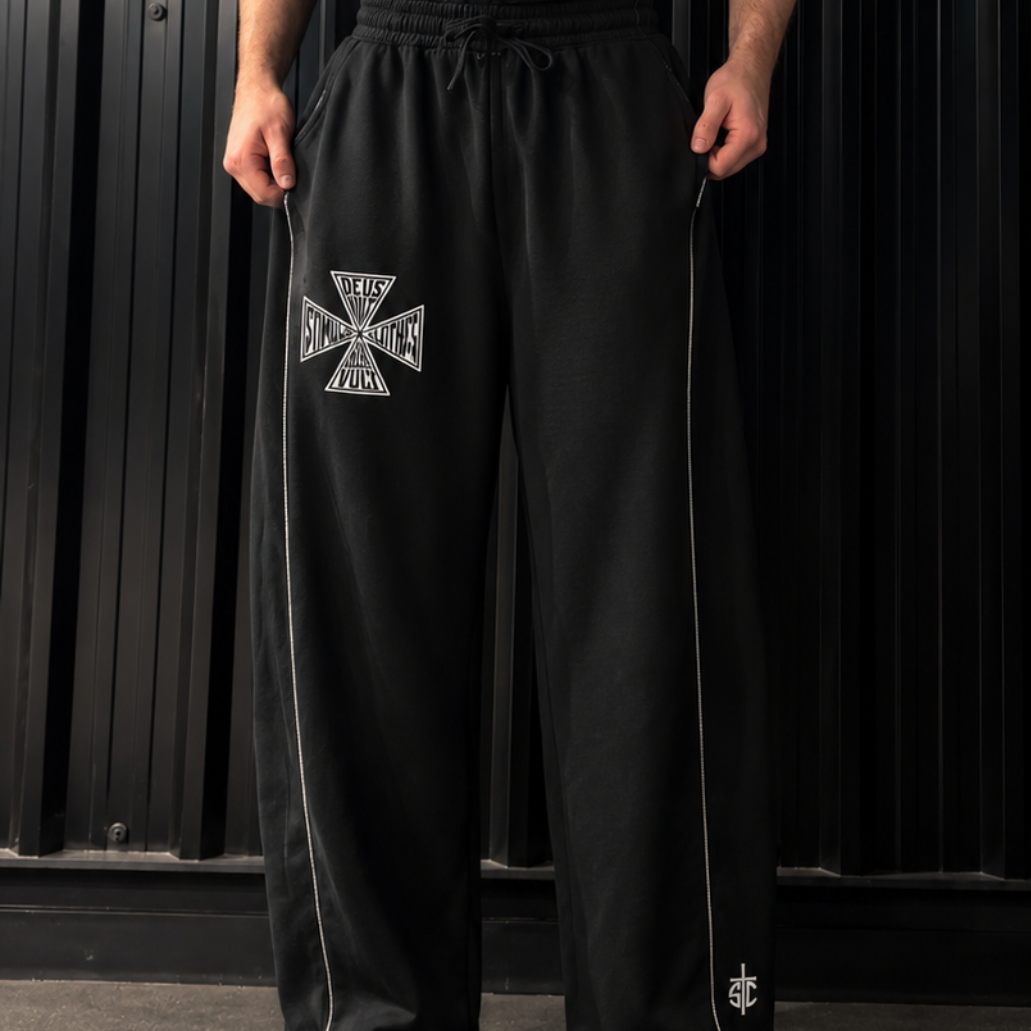 A man wearing a black tank top with 'stimulus' printed on the chest and black sweatpants with a silver Iron Cross emblem on the thigh, standing against a black corrugated metal backdrop.