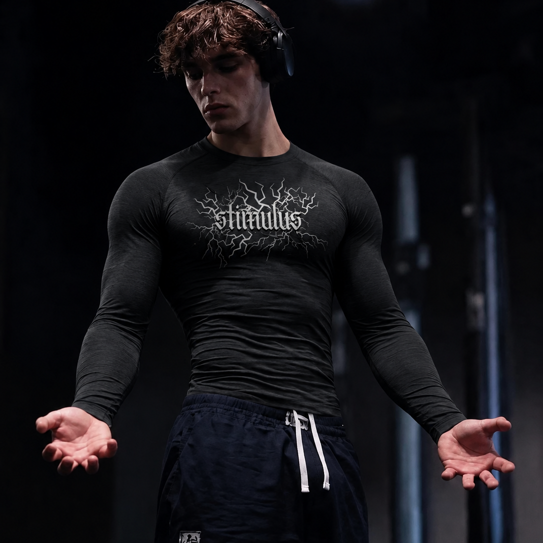 A man wearing a black long-sleeve athletic t-shirt with a white 'stimulus' graphic on the chest, paired with dark navy drawstring sweatpants, standing in a dark gym environment.