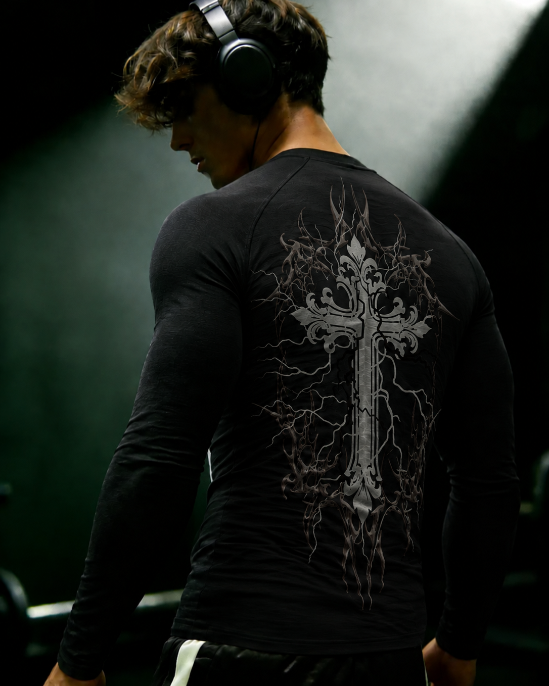 A man wearing a black long-sleeve athletic compression shirt with a large gray gothic cross and lightning bolt graphic on the back, shown from behind in a dark gym setting.