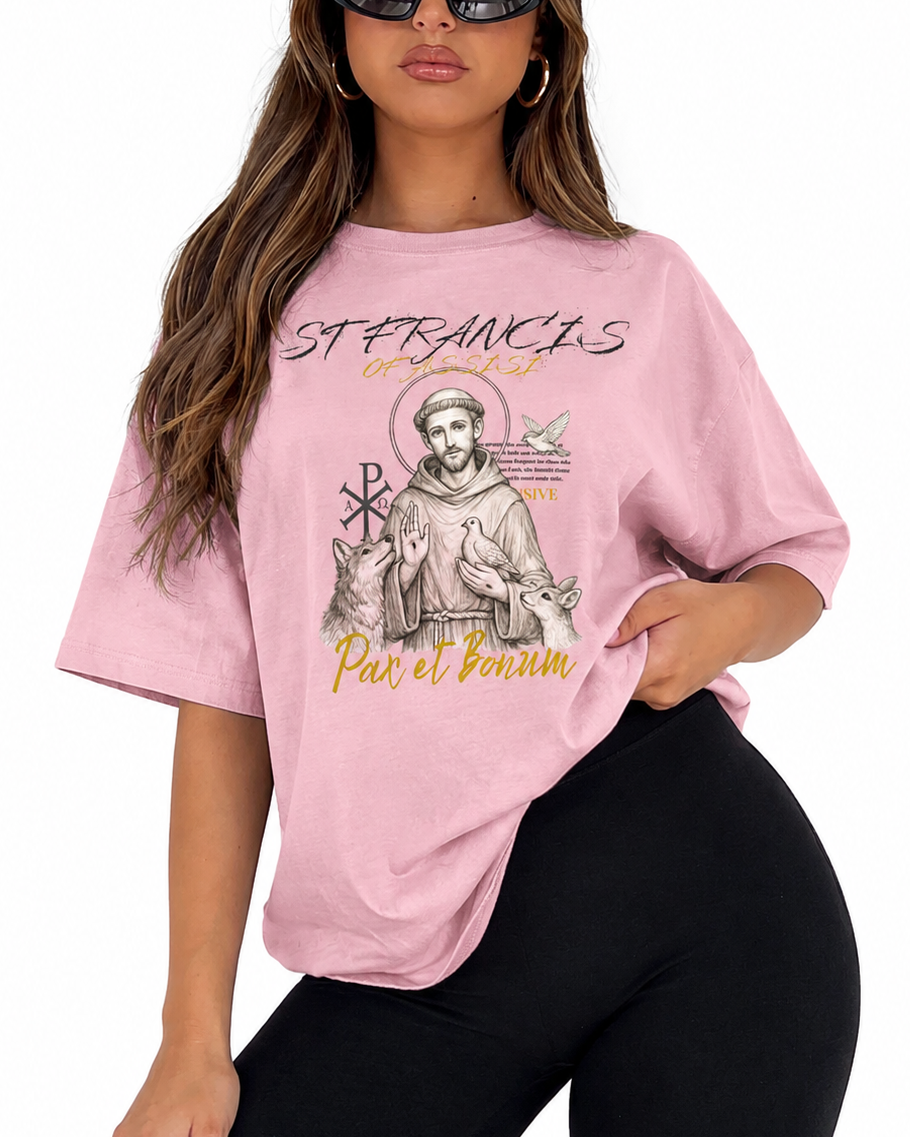 A woman modeling an oversized light pink graphic t-shirt featuring Saint Francis of Assisi with the text 'ST FRANCIS PAES BORUM' and 'Par et Bonorum'. She is wearing black leggings and black sunglasses against a plain white background.
